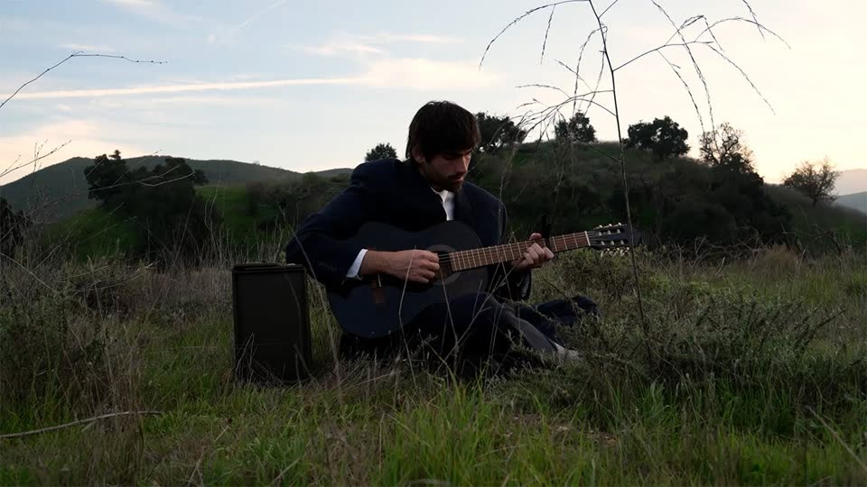 Daniel in a suit playing guitar on a hillside